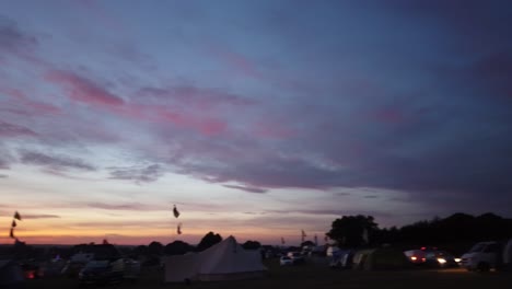 pan de un atardecer naranja, rosa y azul en un festival de música, que contiene un campo de furgonetas de campamento con siluetas, tiendas de campaña, banderas