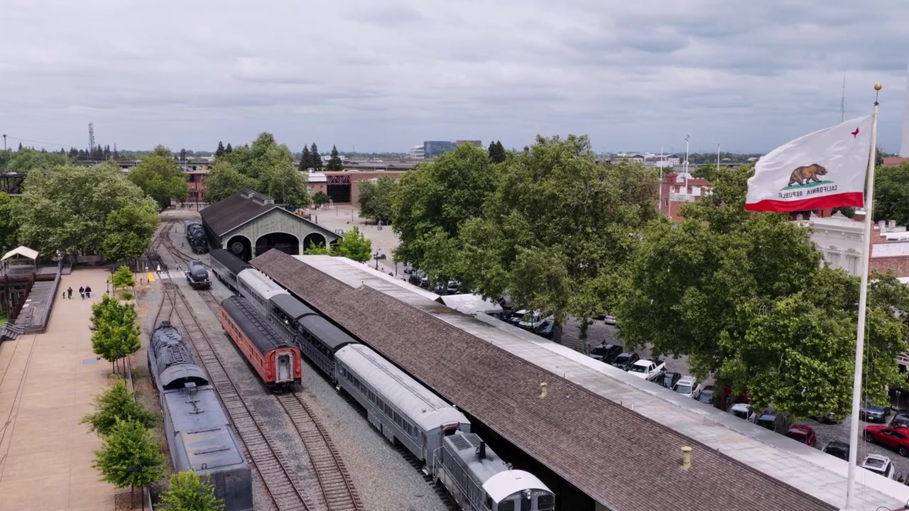 Museo ferroviario storico di Sacramento con treni d'epoca sui binari, una grande bandiera dello stato della California che sventola, piattaforma di deposito coperta e alberi verdi lussureggianti sotto un cielo nuvoloso e coperto. Ripresa aerea in ascesa