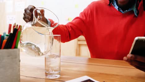 man pouring jug of water in glass while using mobile phone