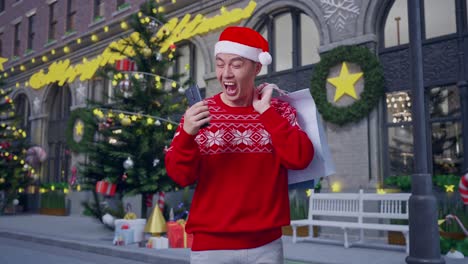 asian man in santa suit with the shopping bags celebrating receiving a discount while using smartphone shopping online in christmas night