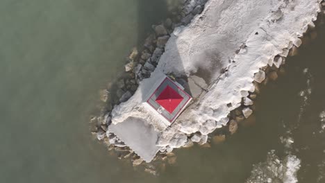 Toma-Aérea-De-Una-Vibrante-Cabaña-Roja-En-Un-Muelle-Nevado-De-Southampton,-Rocas-Visibles-Junto-Al-Agua,-Temporada-De-Invierno,-Estructura-Aislada