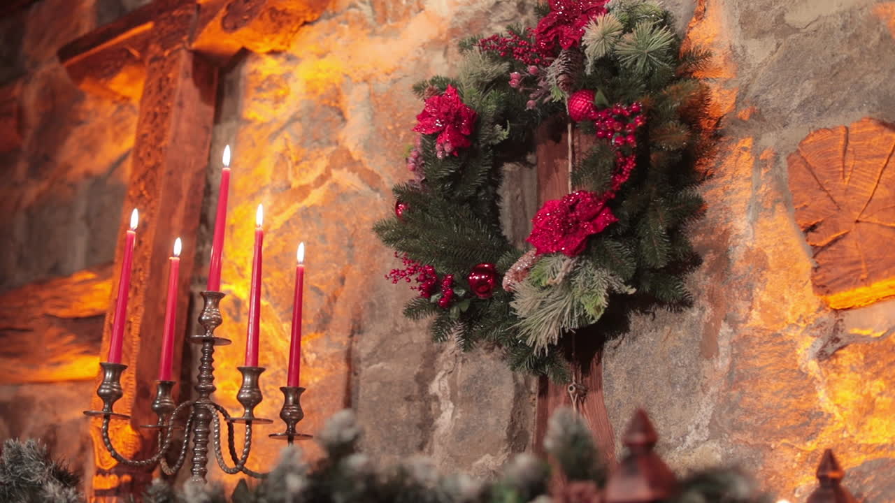 Fir-tree wreath with bright artificial flowers and decorations. Stone wall lit with dim soft light of candles .