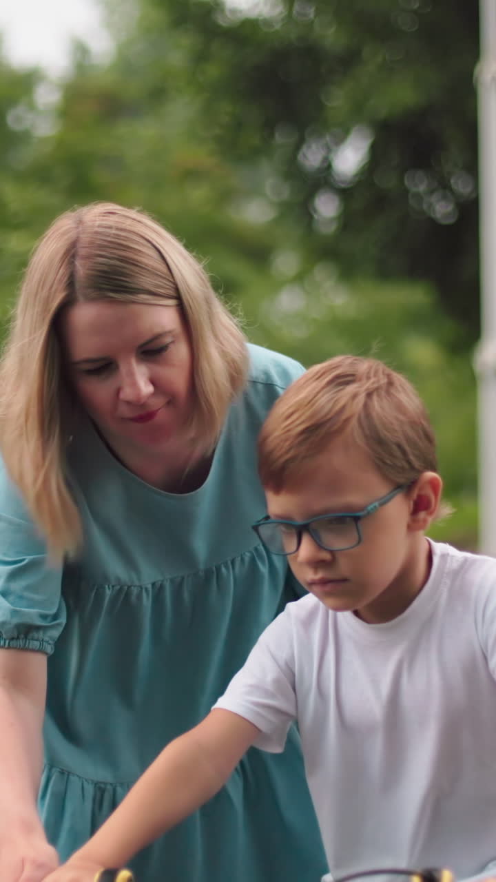 una madre guía a su hijo mientras anda en bicicleta, pero el niño parece infeliz, con vegetación en el fondo