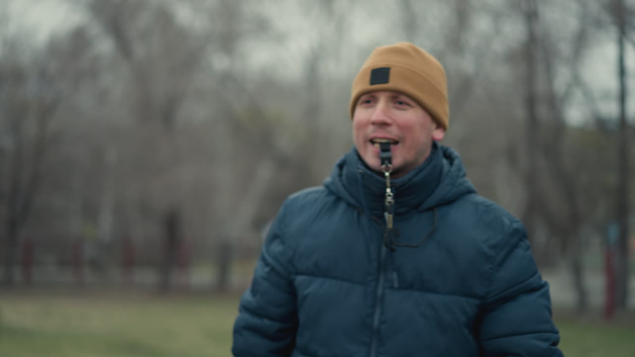 A coach holding a soccer ball in a field with a whistle in his mouth, handing the ball to a young boy wearing glasses and a black beanie, with a blur background