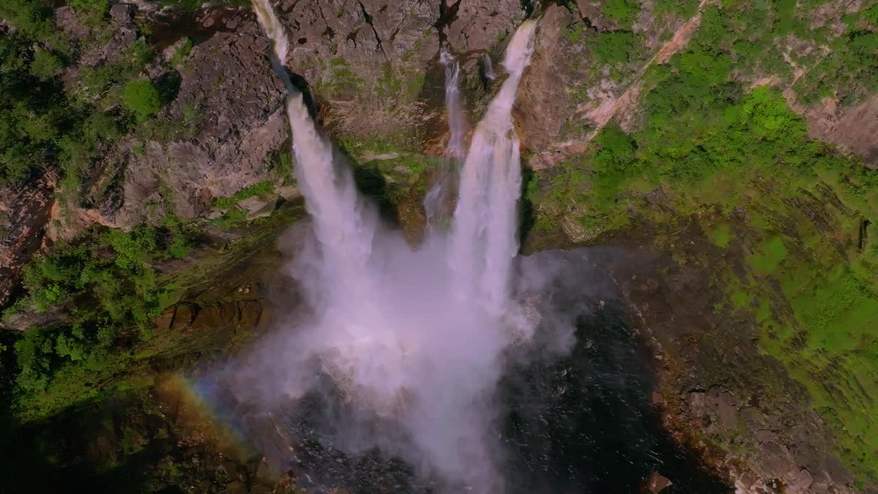 Capturing an aerial view of a stunning waterfall cascading into a rocky gorge, creating a rainbow amidst the lush green cerrado of Chapada Dos Veadeiros National Park, Brazil, slow motion drone shot