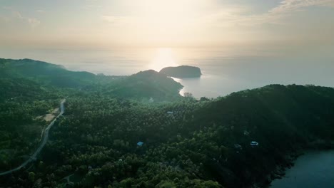 aerial establishing pullback and pan of the coastline of koh phangan with the sun reflecting on the water, surrounded by green forested hills and beaches