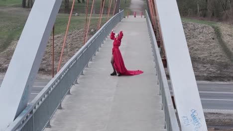 Smooth-aerial-drone-shot-of-a-beautiful-model-with-bright-red-dress-on-a-bridge-spinning