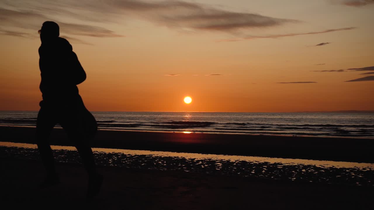 日没時に背中の砂浜でギターを持って走っている男