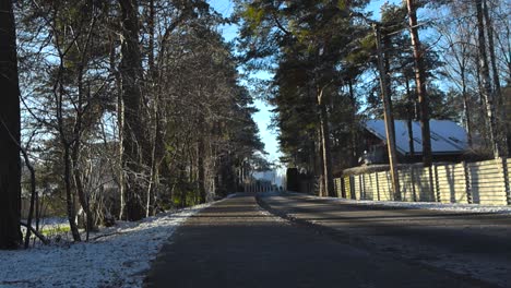 a lonely asphalt road during winter time while sun is shining and fences are on the sides. a pedestrian road also on the side of the road. snow covering the ground while sun shines through the trees.