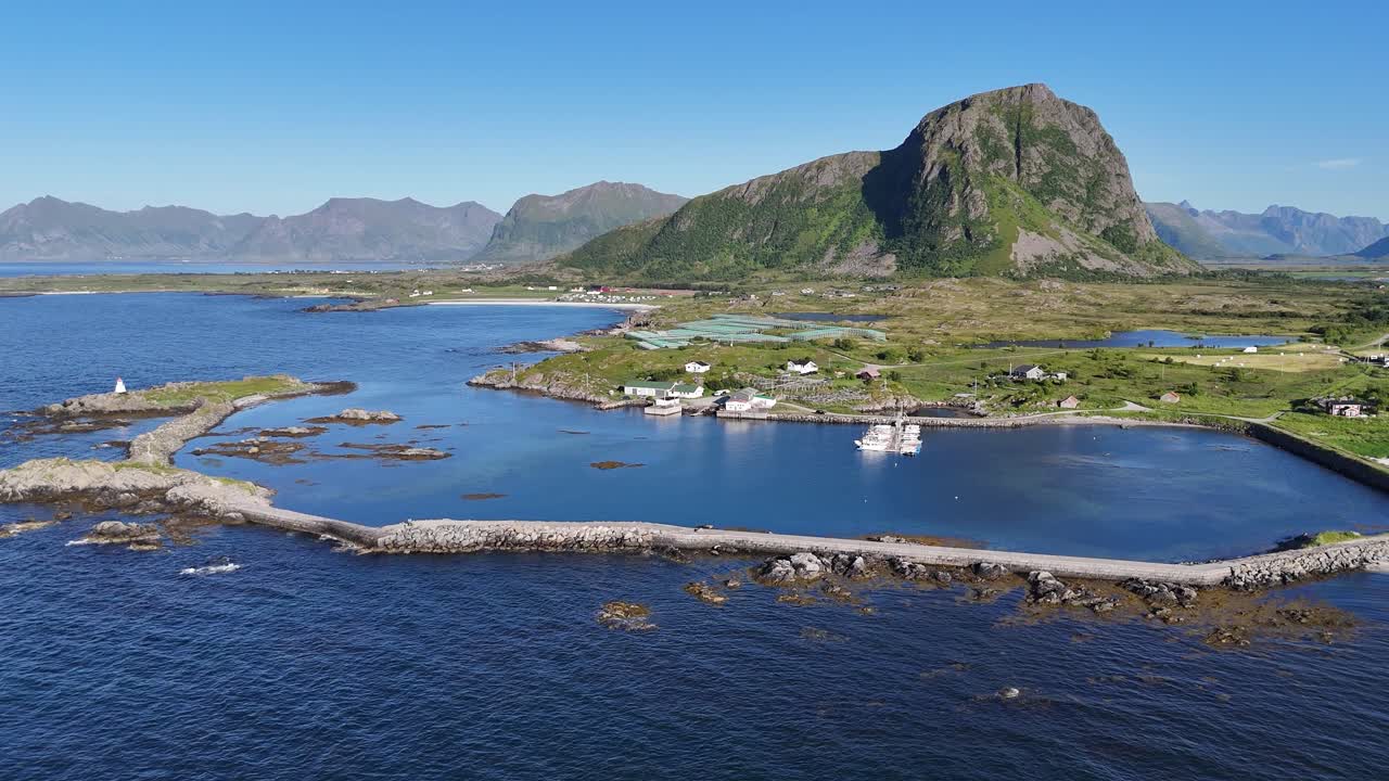 Aerial view of Gimsøy Island in Lofoten, Norway. The concrete pier extends into calm blue water, surrounded by small boats, green fields, and scattered white Norwegian houses