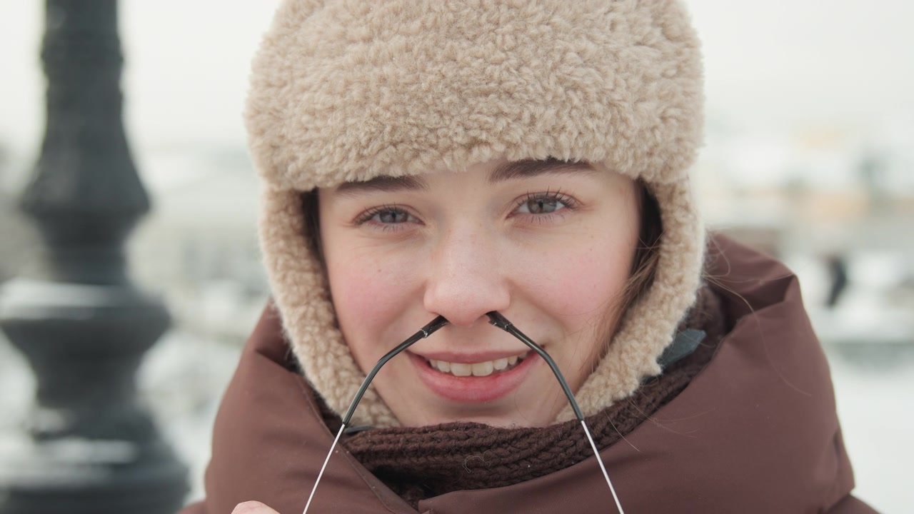 Close-up head-and-shoulders portrait of young person in fluffy fur-lined trapper hat, padded brown coat, and layered knit scarf, smiling while lowering metal-framed sunglasses and holding frames