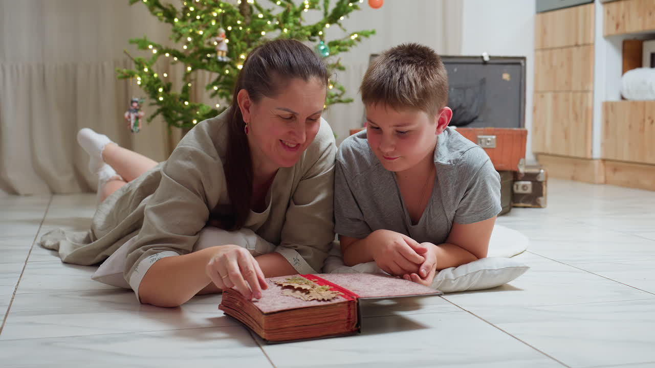 mom and son in casual clothes lying on marble floor opening large vintage book smiling warmly cozy festive indoor setting background featuring christmas tree with lights family moment together