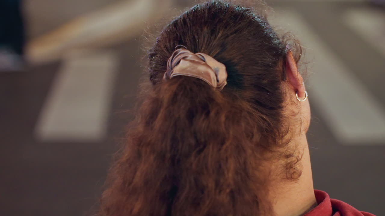 Giovane donna all'attraversamento pedonale con gli auricolari sotto i lampioni, che indossa occhiali e uno scrunchie, mentre aggiusta un auricolare e guarda in entrambe le direzioni, un ritratto cinematografico notturno della città che trasmette quieta contemplazione.