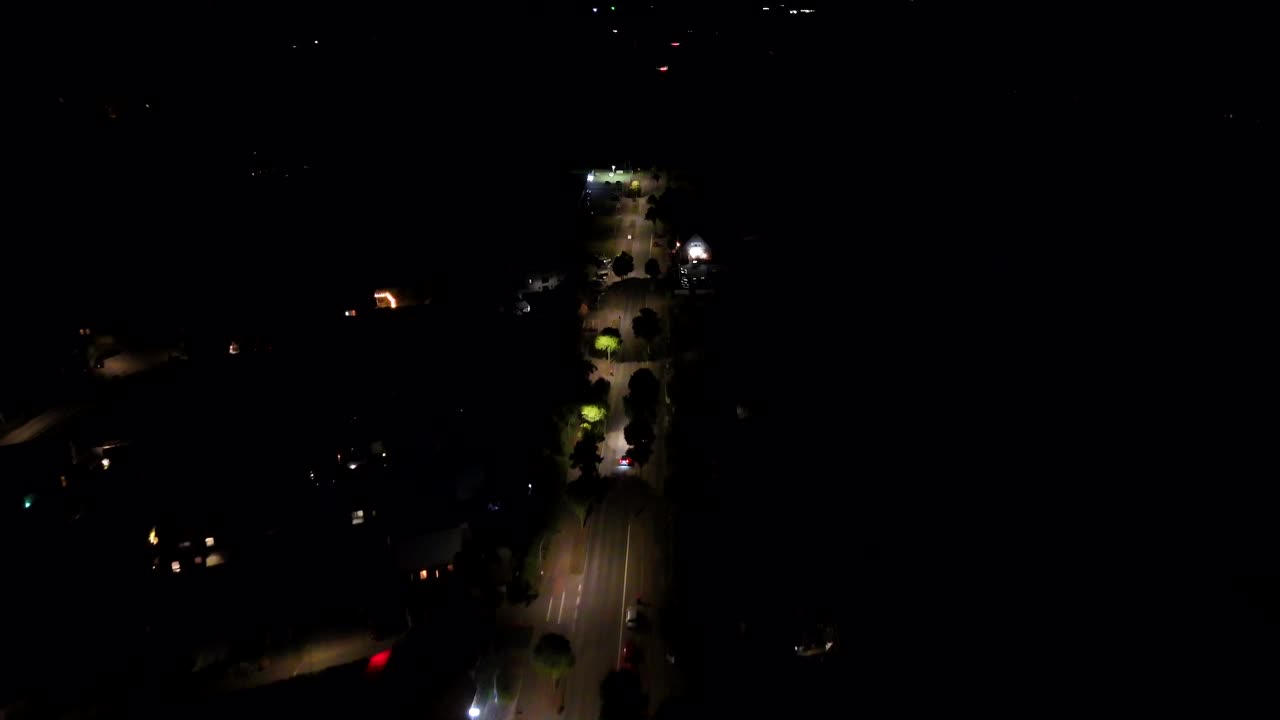 Illuminated street in American town at night. Aerial Birds Eye shot. Glowing lantern and streetlamps on quiet small neighborhood. Driving car on road. Top down