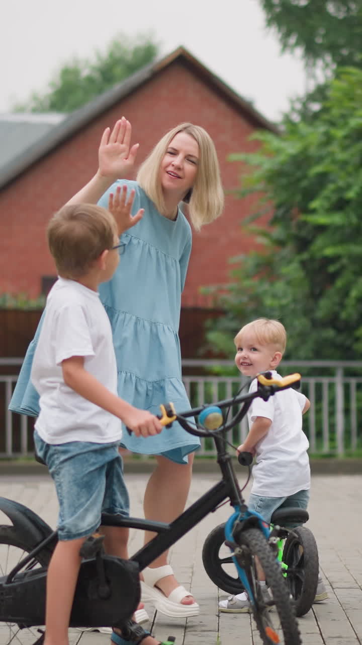 한 어머니가 집 앞의 포장된 길에서 아이들과 대화하는 것을 볼 수 있습니다. 그녀는 손을 고 큰 아이에게 기다리라고 말하면서 아이에게 하이 파이브를합니다.