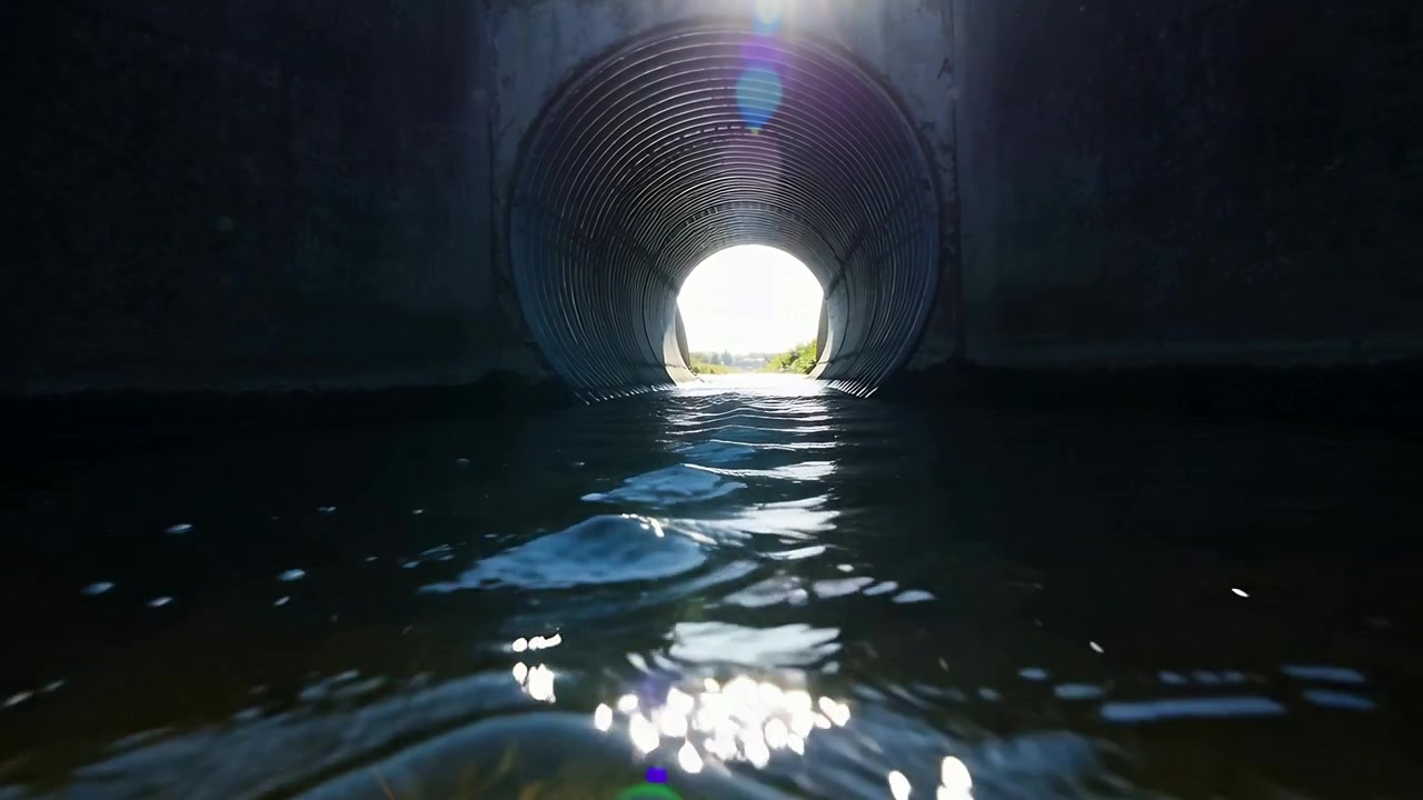Plätscherndes Wasser, das auf Sonnenlicht reagiert, in einem Durchlass und flachen Bach, bewegt sich in einem Metallrohr auf die Linse zu.