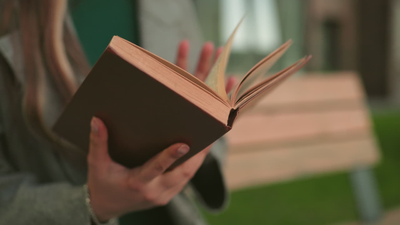 Close up of woman holding open book outdoors while standing near wooden bench, fingers gripping pages with natural light, detail view highlighting reading moment and lifestyle atmosphere