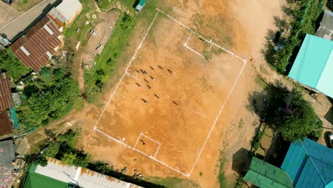 4K-Cinematic-sport-aerial-drone-footage-of-children-playing-on-a-footbal-field-in-the-mountain-village-of-Doi-Pui-next-to-Chiang-Mai,-Thailand-on-a-sunny-day