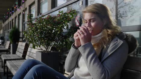 Woman-blows-on-and-sips-a-cup-of-hot-coffee,-sitting-on-a-street-bench-in-the-morning