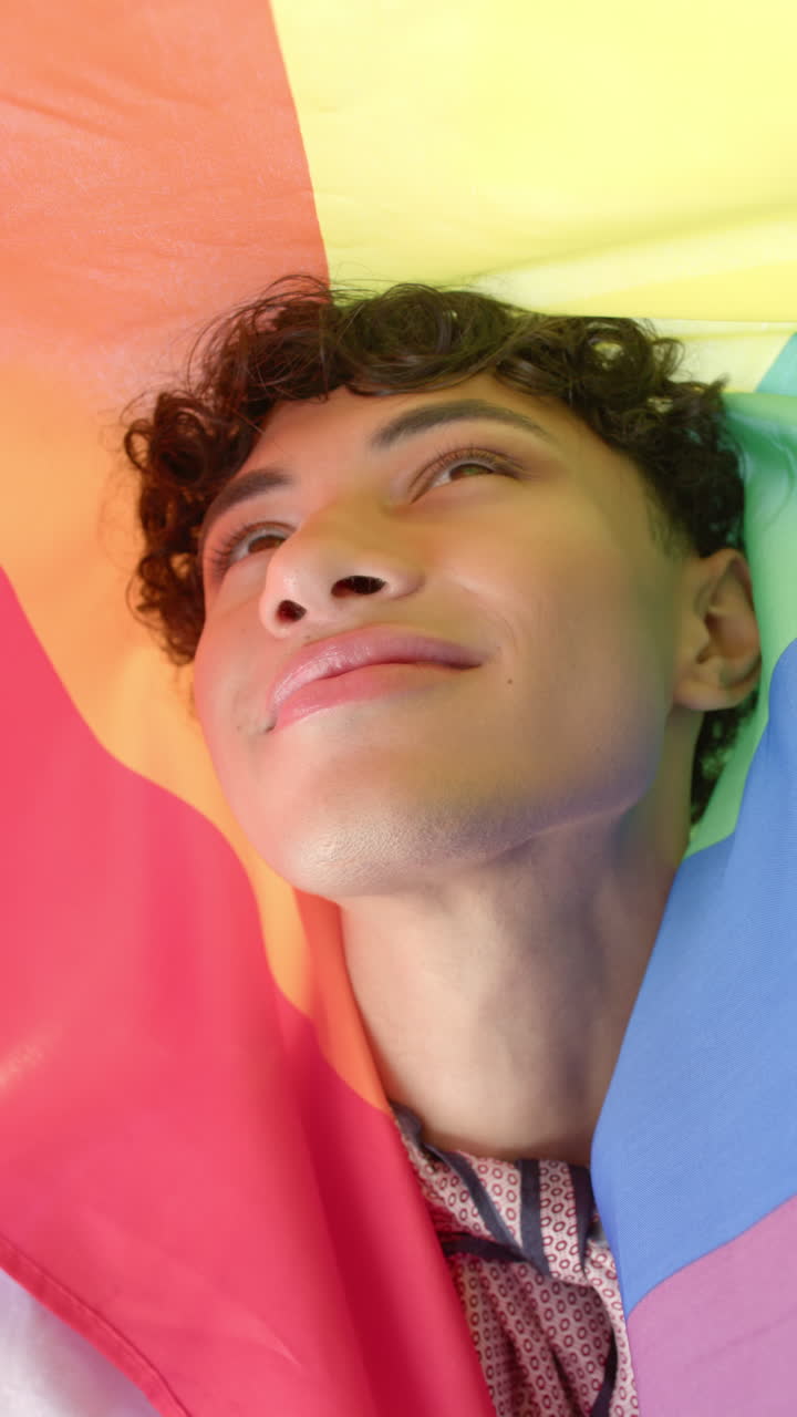 Vertical video: Smiling person wrapped in rainbow flag, embracing pride and joy at home