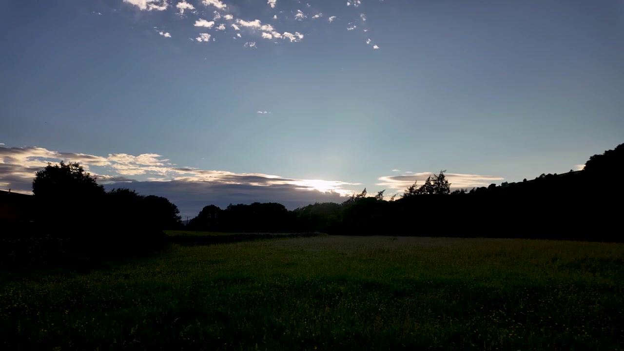 Solnedgangslys synker bak en mørk trelinje mens skyer beveger seg raskt over et bredt landlig felt i timelapse