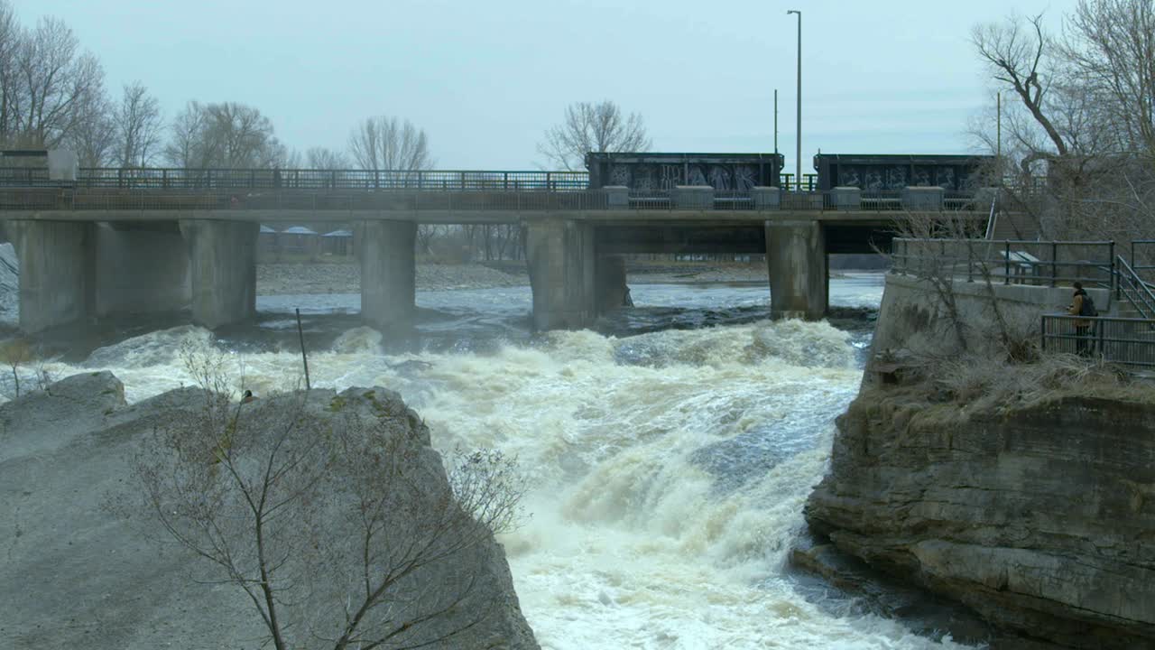 Water pours under a bridge on a river swollen with spring run off as a firetruck passes by on the bridge. Slow Motion