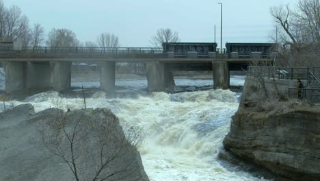 water pours under a bridge on a river swollen with spring run off as a firetruck passes by on the bridge. slow motion