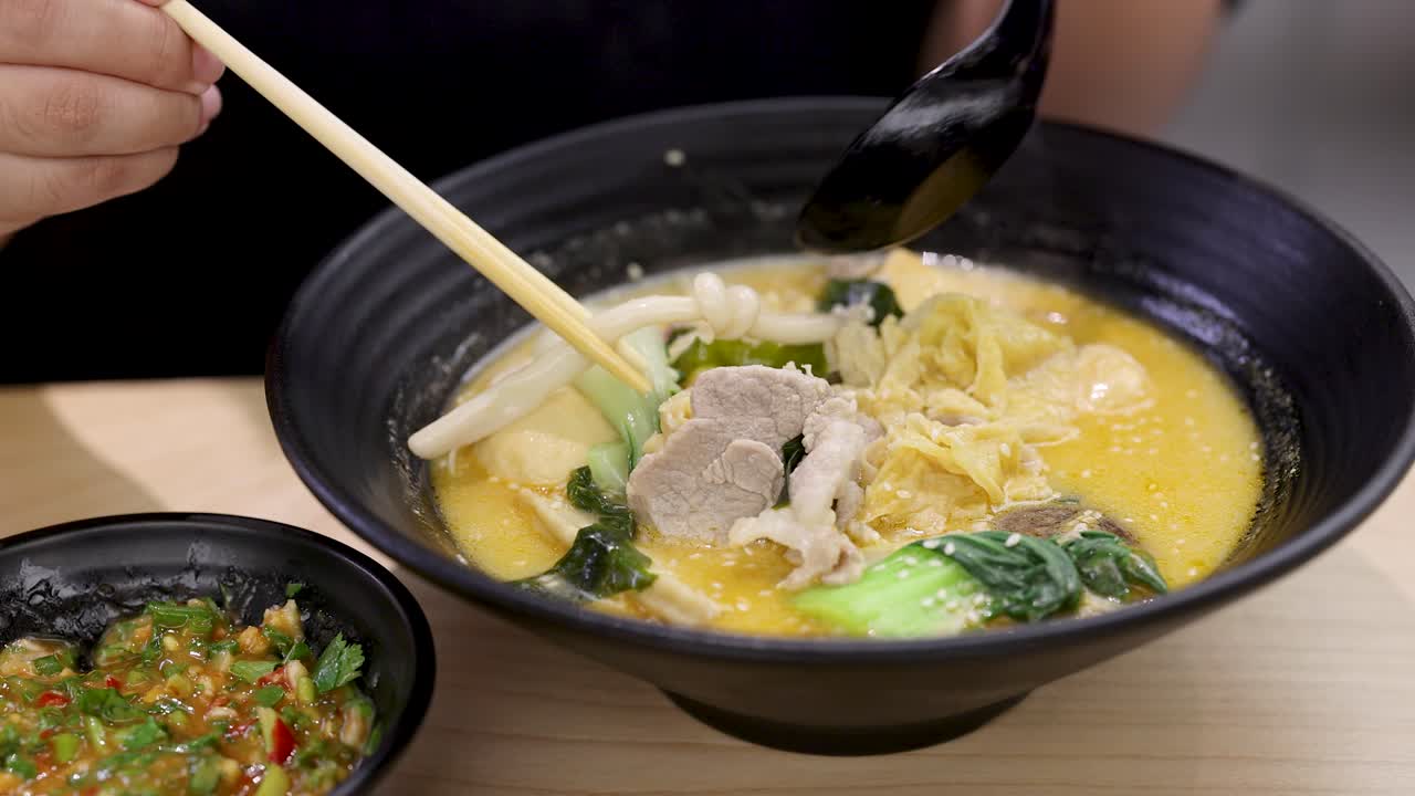 Hand uses chopsticks and spoon to eat spicy noodle soup with vegetables and chicken slices