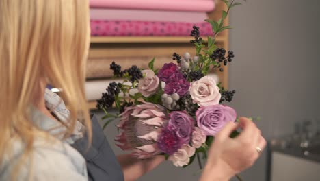 Side-view-of-female-blonde-florist-arranging-modern-bouquet-and-looking-how-the-flowers-are-combined.-Handsome-flower-shot-owner