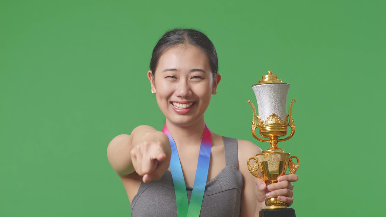 primer plano de una mujer asiática con una medalla de oro y un trofeo tocando su pecho luego sonriendo y señalando a la cámara estando orgullosa de sí misma en el fondo de la pantalla verde en el estudio