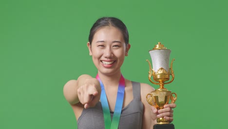 close up of asian woman with a gold medal and trophy touching her chest then smiling and pointing to camera being proud of herself on green screen background in the studio