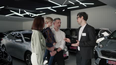 a confident young brunette man in a business suit and glasses an assistant in a car dealership tells a man to his wife and little son about what cars are and about them