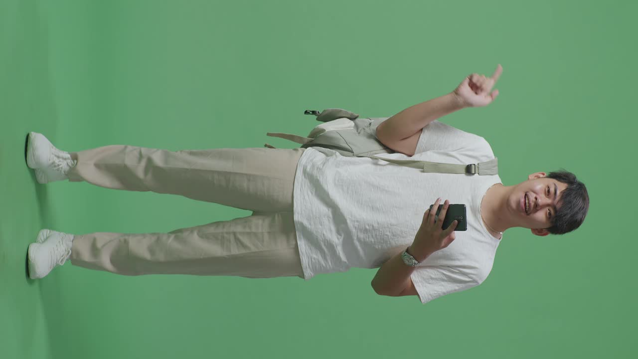 cuerpo lleno de estudiante hombre asiático usando teléfono móvil y dedos directos lado en pantalla verde estudio
