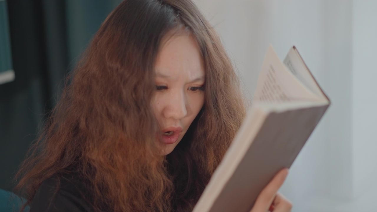 Medium close-up of young woman leaning forward while reading from open book, lips forming words and eyes fixed on pages indicating active vocalization or surprised reaction; hands gripping book