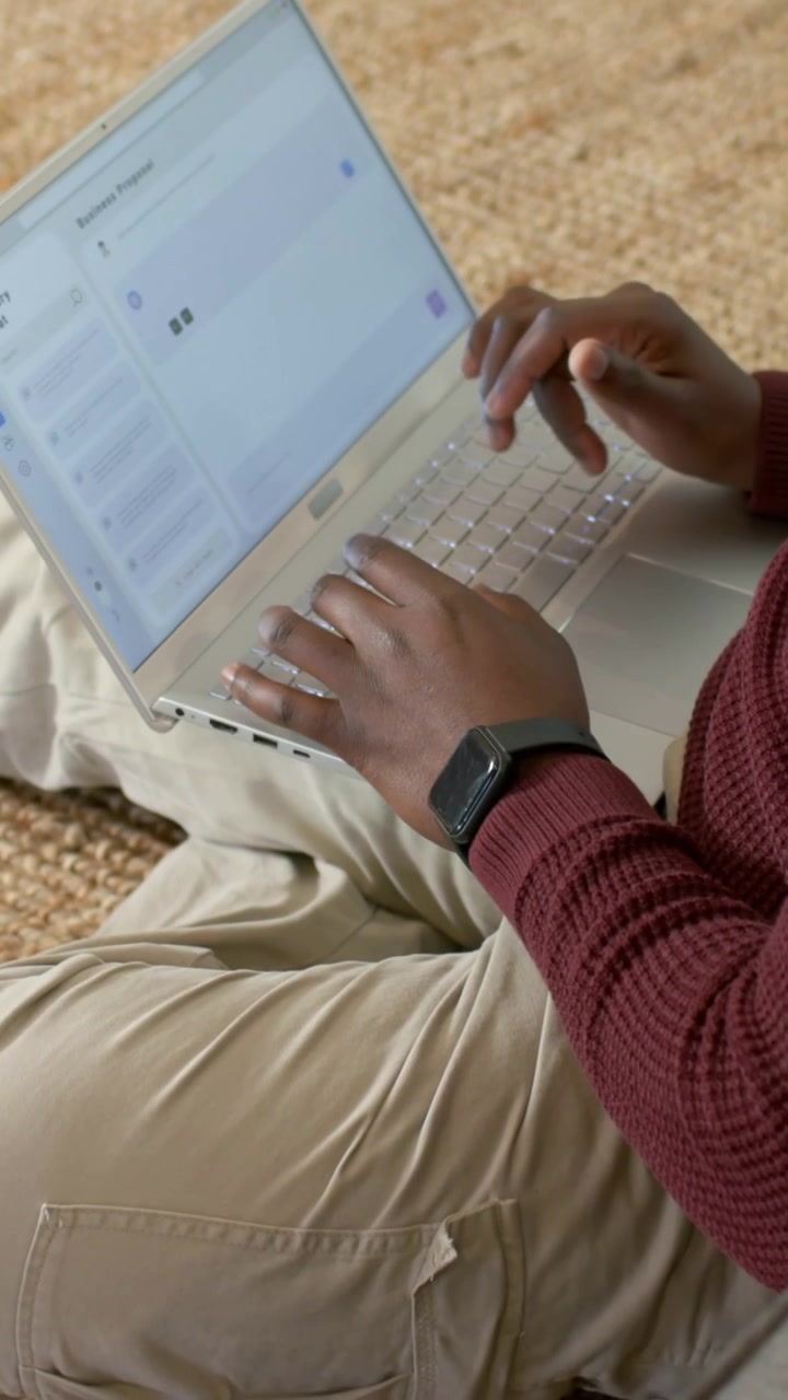 Vertical video: Typing man in maroon top, hands settling, composing on laptop on carpet with watch
