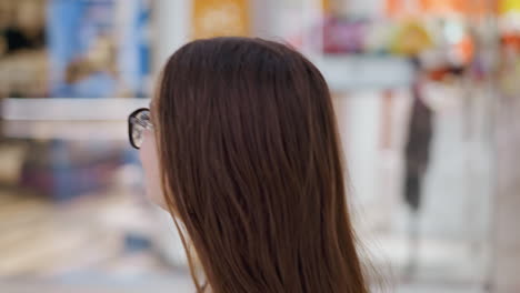 안경을 입고 긴 머리카락을 가진 여성이 부드럽게 조명 된 소매 공간에서 앞으로 바라보고있는 클로즈업 측면 뷰, 보케 조명과 함께 따뜻한 톤의 흐릿한 상점 인테리어는 현대적인 쇼핑 분위기를 만니다.