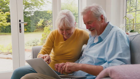 retired senior couple sitting on sofa at home using laptop online shopping or booking holiday