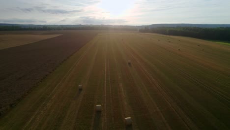 Drone-Aéreo-Hacia-Adelante-Toma-De-Luz-Solar-Que-Cae-Sobre-Un-Campo-Dorado-Con-Fardos-De-Heno-O-Fardos-De-Paja-Después-De-La-Cosecha