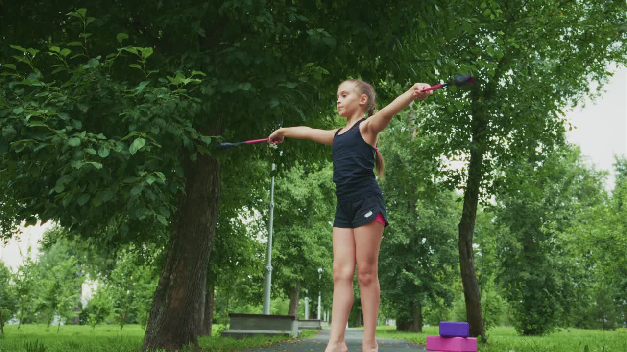 A young girl demonstrates impressive balance and poise while practicing rhythmic gymnastics in a serene park setting, showcasing her talent with rhythmic apparatus