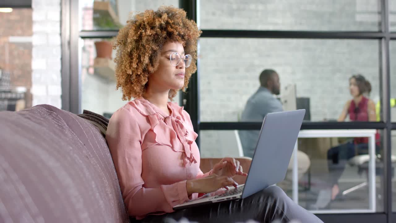 Thoughtful biracial casual businesswoman using laptop on couch in office, copy space, slow motion