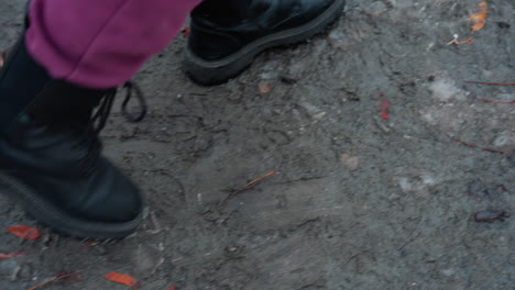 close-up of lower legs in maroon trousers and black boots walking on a worn, icy path covered with scattered foliage and rough, patchy terrain, footsteps leave imprints on frozen ground