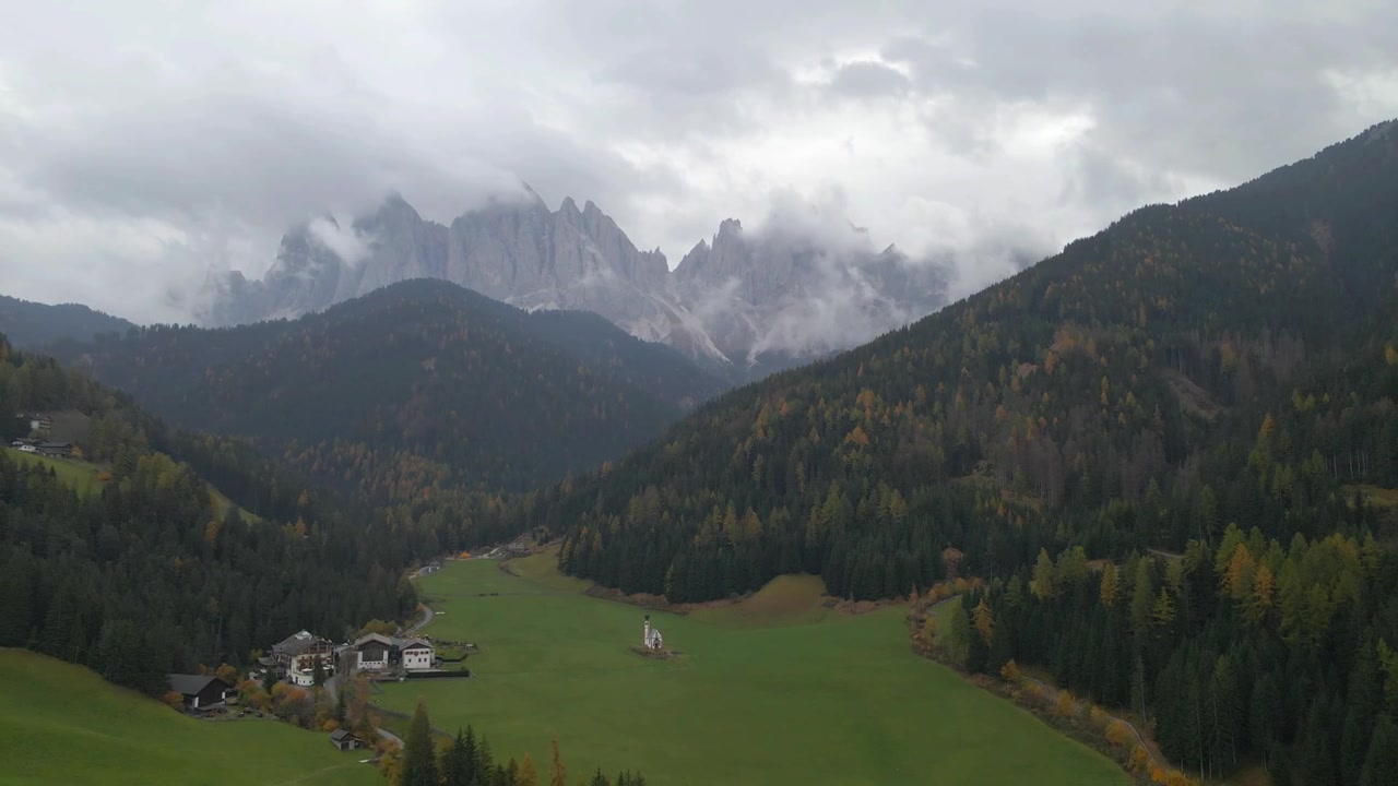 Immersa tra prati rigogliosi e fitte foreste, la chiesa e il villaggio di Santa Maddalena in Val di Funes mostrano la maestosa catena montuosa delle Odle, Alto Adige, Italia, con drone in allontanamento.