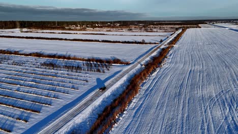 Escena-Aérea-De-Un-Automóvil-Rural-Con-Un-Vehículo-Solitario-Conduciendo-Por-Una-Carretera-Cubierta-De-Nieve-Que-Serpentea-A-Través-De-Un-Campo,-Con-Una-Línea-De-árboles-Oscuros-En-La-Distancia-Y-Un-Cielo-Azul-Claro-En-Lo-Alto,-Toma-Panorámica-En-órbita