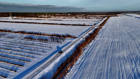 aerial rural car scene with a lone vehicle driving on a snow covered road that winds through a field, with a line of dark trees in the distance and a clear blue sky overhead, orbit pan shot