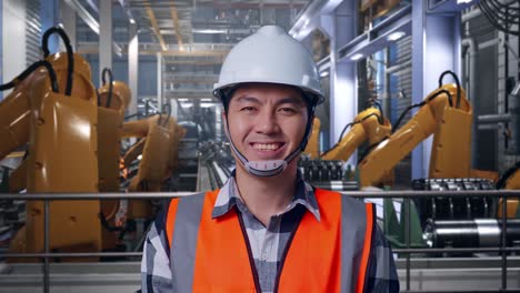 close up of asian male engineer with safety helmet smiling to camera while standing with industrial robotic arms working