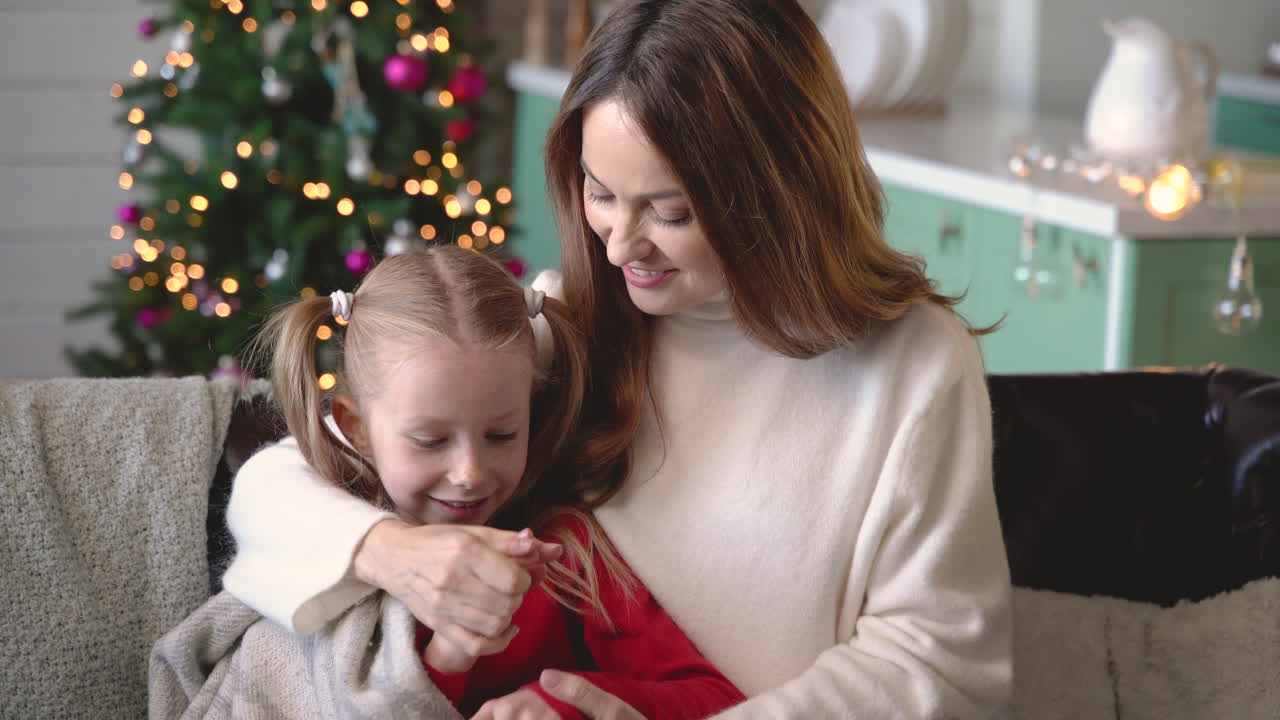 madre e hija sentadas en el sofá cubiertas por una manta mientras hablan en la sala de estar con adornos navideños