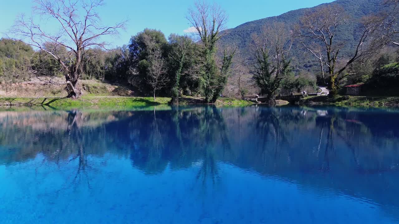 Voo cinematográfico de drone em baixa altitude sobre as nascentes turquesa do rio Louros. Vista de cima da água azul-profundo e árvores exuberantes refletindo na superfície espelhada. Natureza de água doce deslumbrante