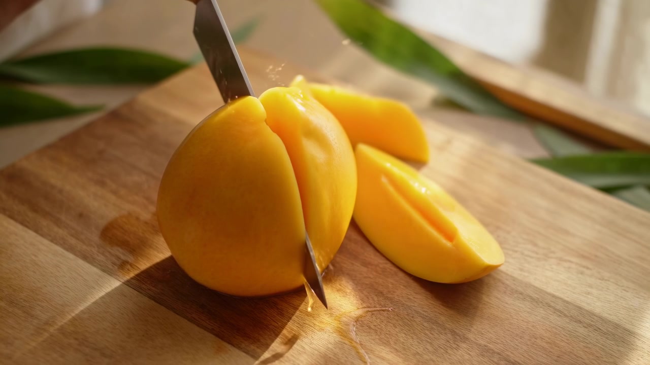 Primo piano di un coltello che taglia un mango giallo maturo su un tagliere di legno, rivelando segmenti lucidi, goccioline di succo fresco e una consistenza ricca, il tutto sotto una luce calda e in un'atmosfera vivace di preparazione in cucina, con mango e coltello.