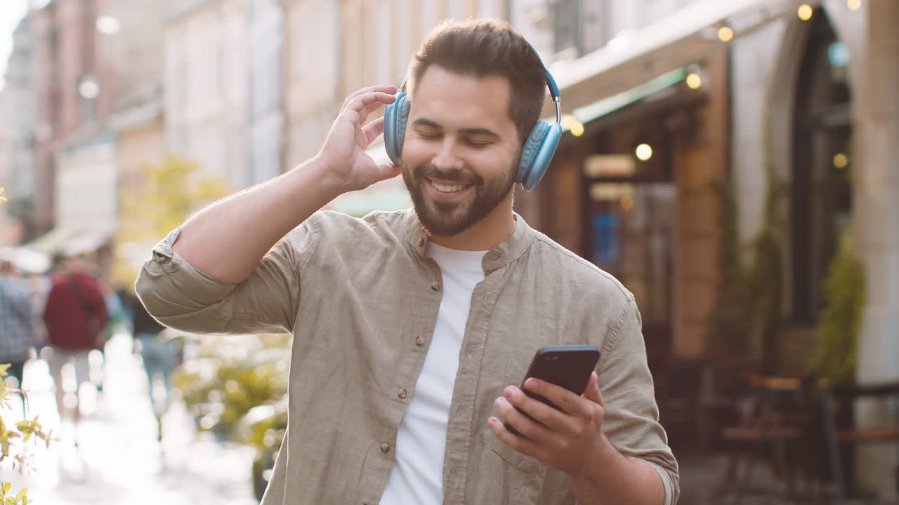 Giovane uomo felice con cuffie wireless che sceglie e ascolta musica, ballando all'aperto in una strada cittadina.