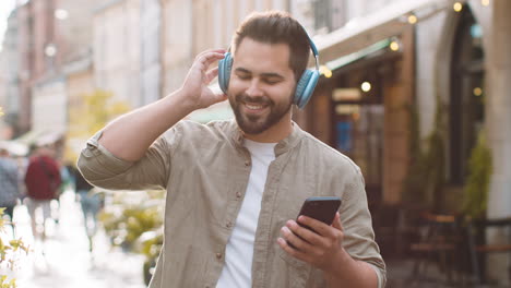 Happy-young-man-in-wireless-headphones-choosing,-listening-music-dancing-outdoors-city-street